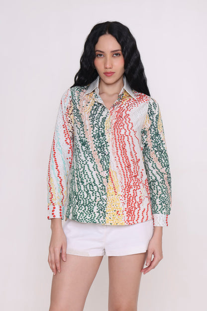A woman modeling a long-sleeve button-up shirt with a colorful vertical dotted stripe pattern in green, red, yellow, and white, paired with white shorts against a plain light background.