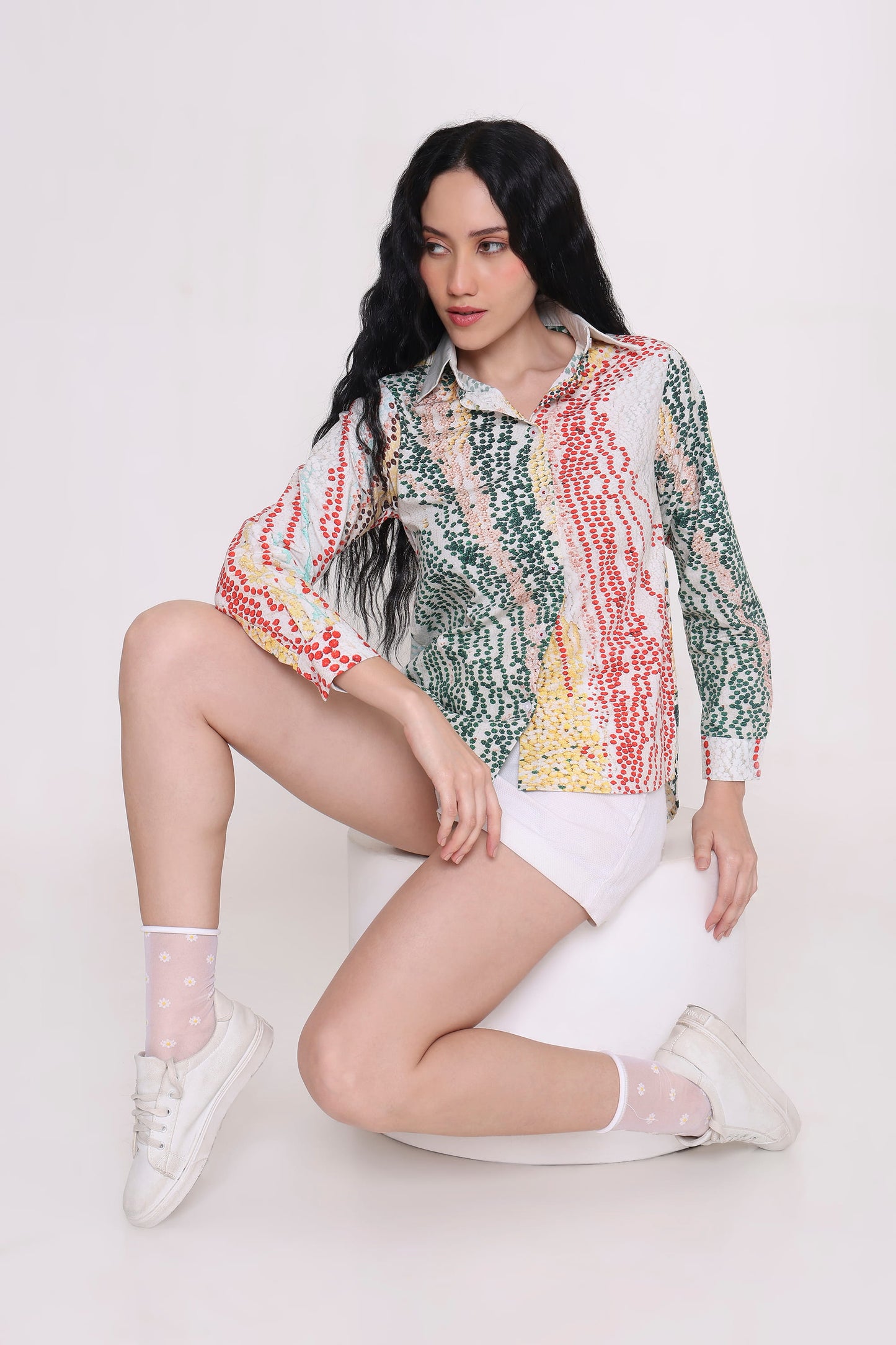 A woman with long black hair sits on a white cylindrical stool against a light gray studio background, wearing a long-sleeve button-up shirt with a multicolor vertical abstract pattern in green, red, yellow, and white, paired with white shorts, white socks, and white sneakers.