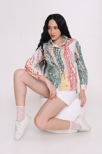 A woman with long black hair sits on a white cylindrical stool against a light gray studio background, wearing a long-sleeve button-up shirt with a multicolor vertical abstract pattern in green, red, yellow, and white, paired with white shorts, white socks, and white sneakers.