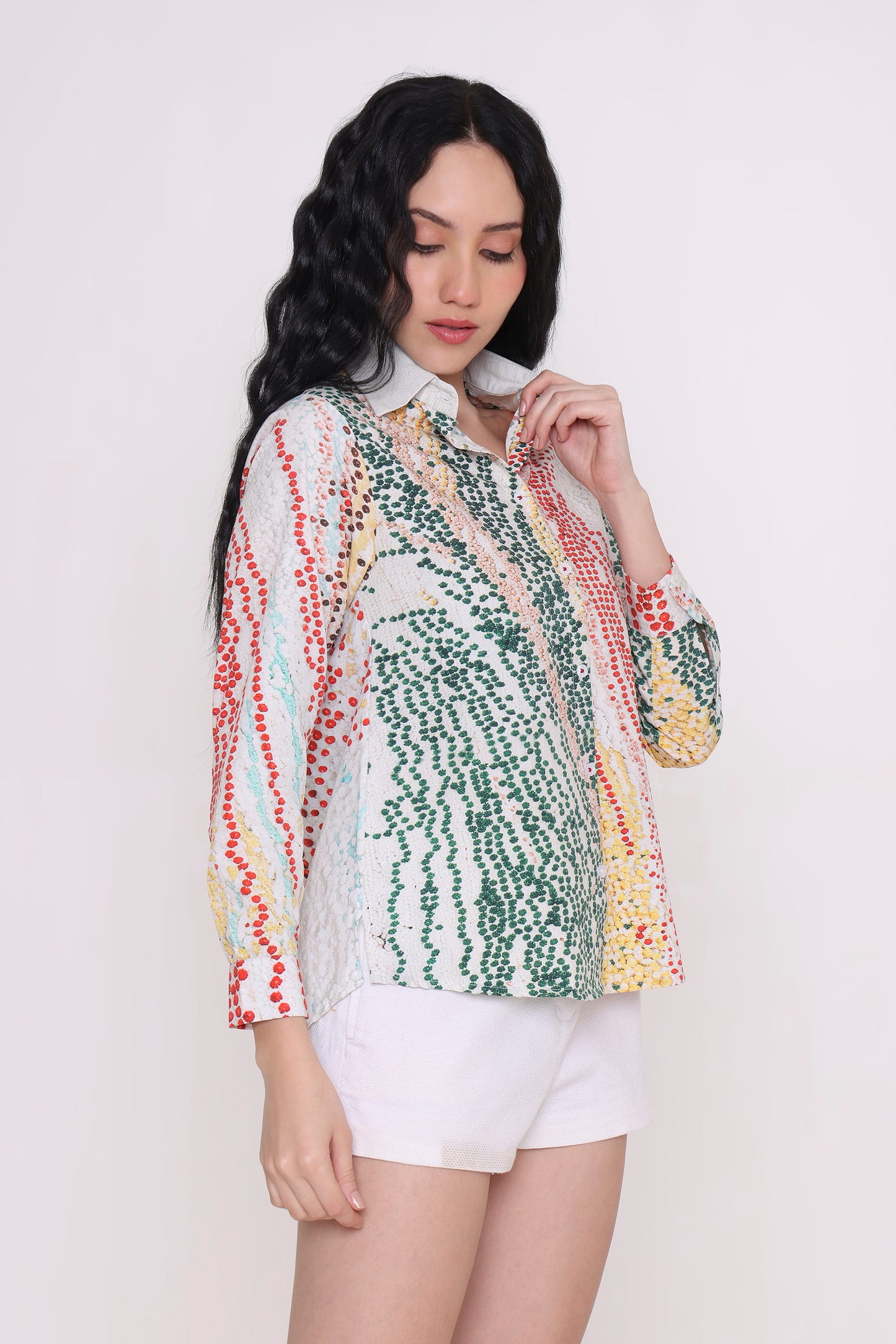 A woman modeling a long-sleeve button-up blouse with a multicolor abstract pattern of green, red, yellow, and blue on a white base, paired with white shorts against a plain light background.
