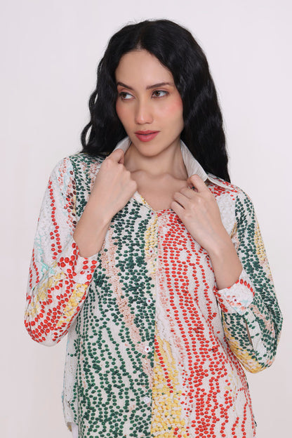A woman with long black hair wearing a long-sleeve button-up shirt with a colorful vertical dotted pattern in green, red, yellow, and white, posing against a plain light background.