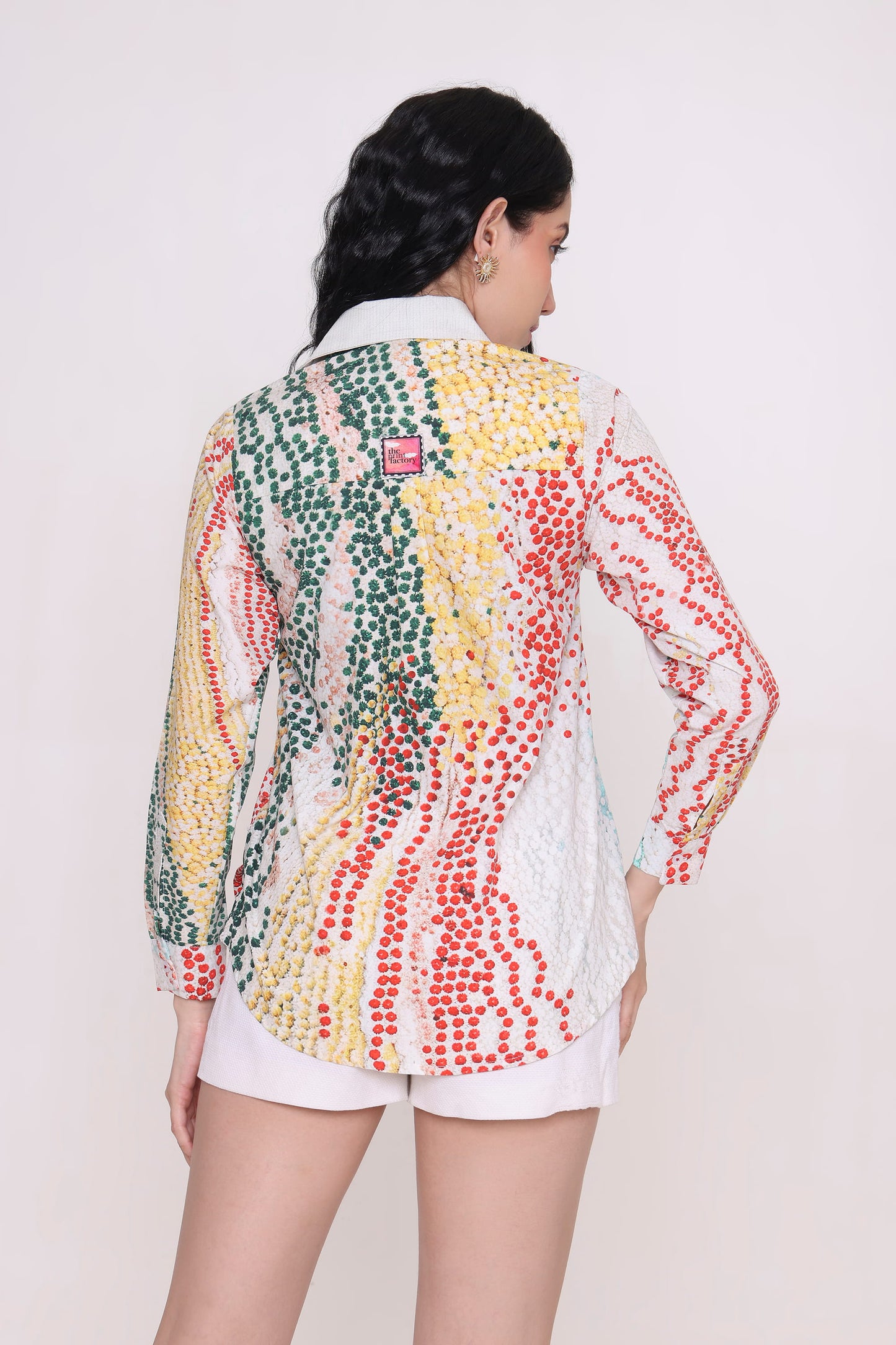 A woman shown from the back wearing a long-sleeve button-up shirt with a colorful vertical dotted pattern in green, yellow, red, and white, paired with white shorts against a plain light background.