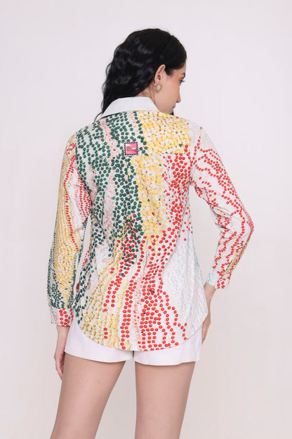 A woman shown from the back wearing a long-sleeve button-up shirt with a colorful vertical dotted pattern in green, yellow, red, and white, paired with white shorts against a plain light background.