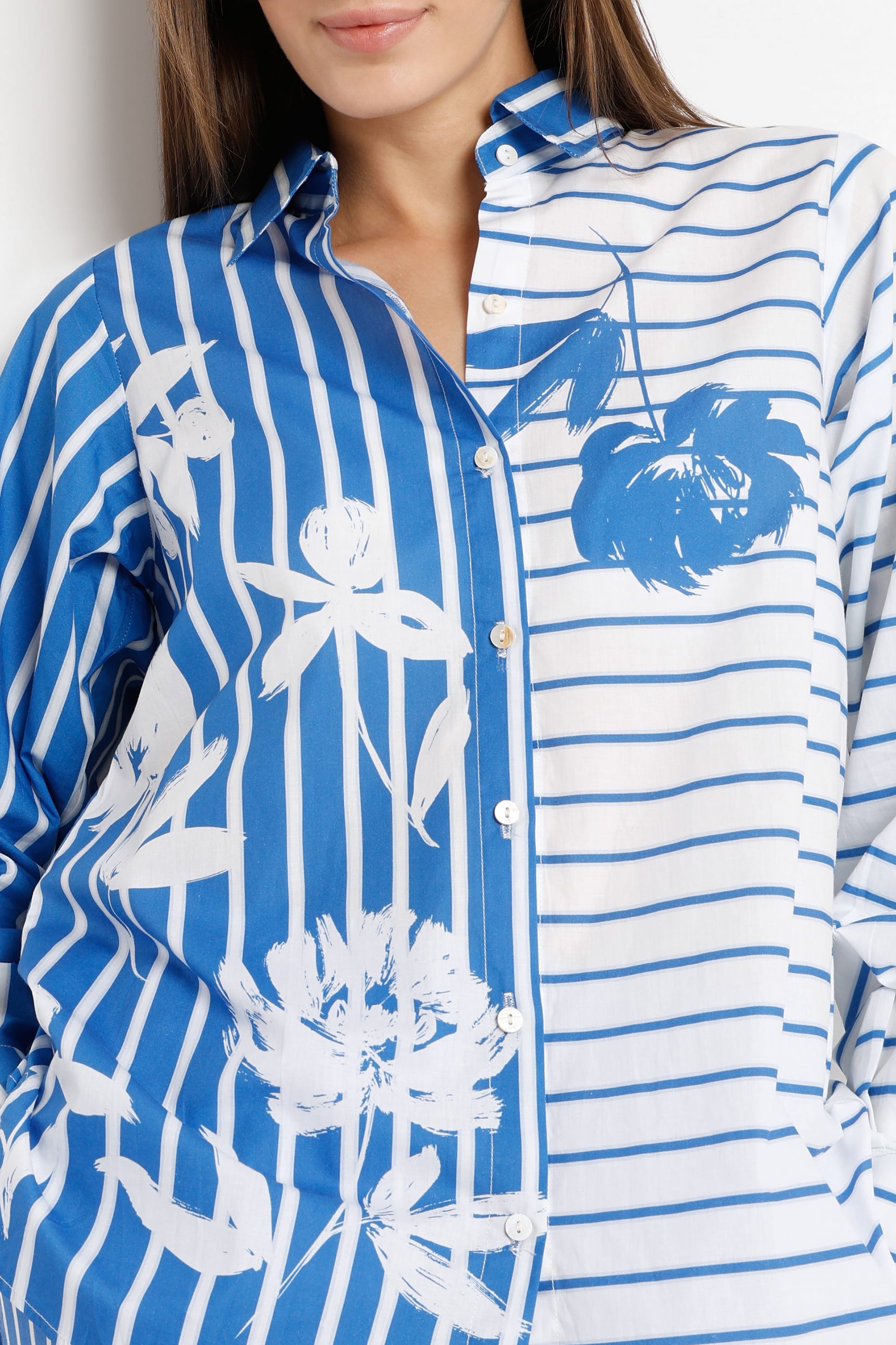A close-up of a woman wearing a blue and white button-up shirt with a split design: one side has vertical blue stripes with white floral motifs, and the other has horizontal blue stripes. The model's lower face and hair are partially visible.
