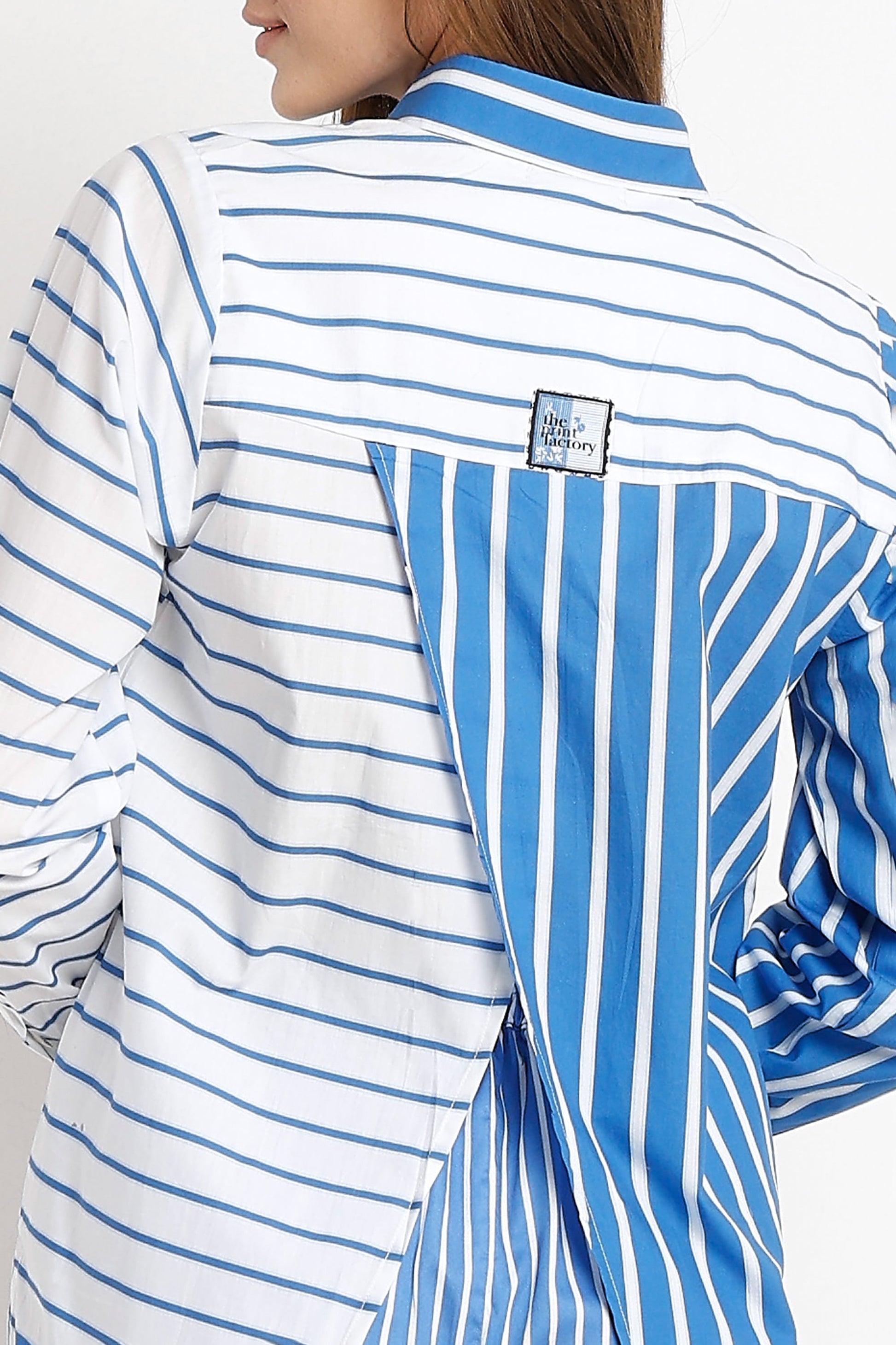 A close-up back view of a model wearing a long-sleeve collared shirt with a split design: one half has thin horizontal blue stripes on white, and the other half has bold vertical blue stripes on white. A small square brand patch is visible on the upper back.