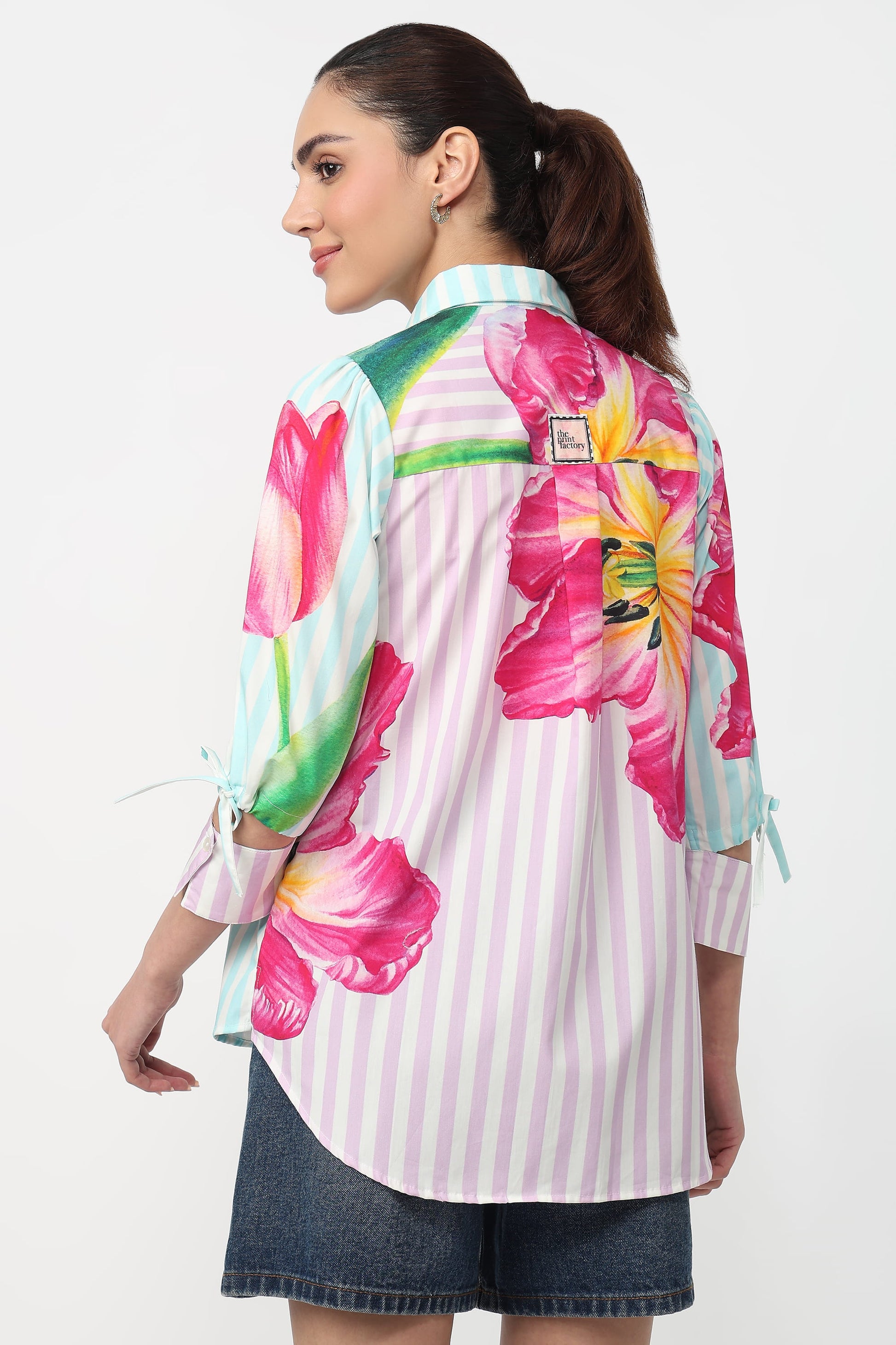 A woman shown from the back wearing a pastel pink and white striped button-up shirt with large pink and yellow floral prints on the shoulders and sleeves, paired with denim shorts against a plain white background.