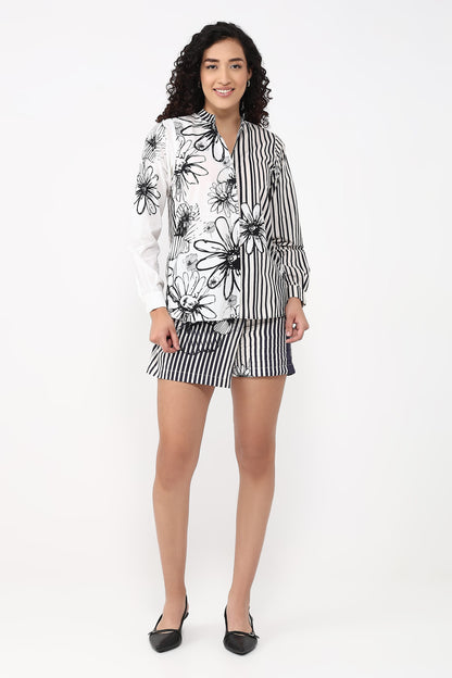 A woman modeling a matching two-piece lounge set featuring a long-sleeve button-up shirt with a black floral print on a white base and striped sleeves, paired with matching black-and-white striped shorts.
