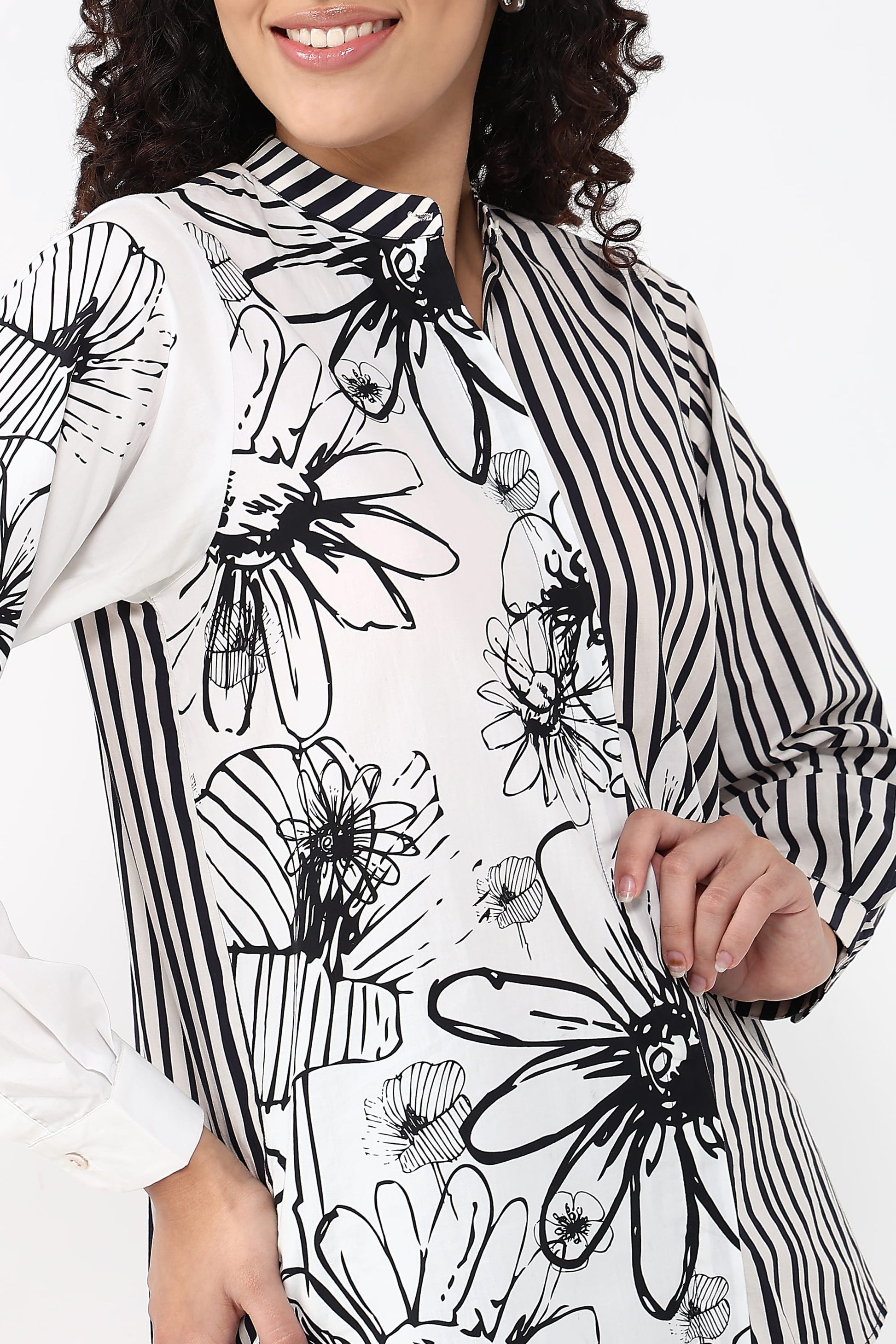 A woman wearing a white long-sleeve tunic top with black floral and striped patterns, shown from the chest up against a plain white background.