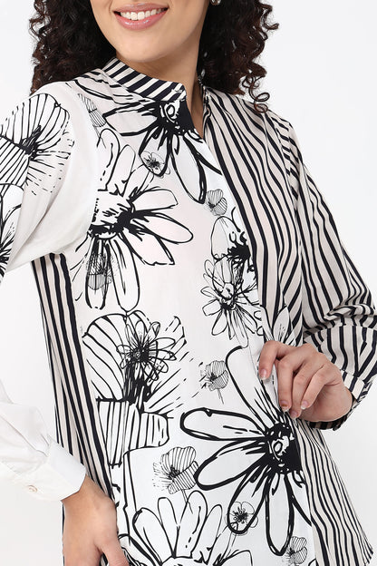A woman wearing a white long-sleeve tunic top with black floral and striped patterns, shown from the chest up against a plain white background.