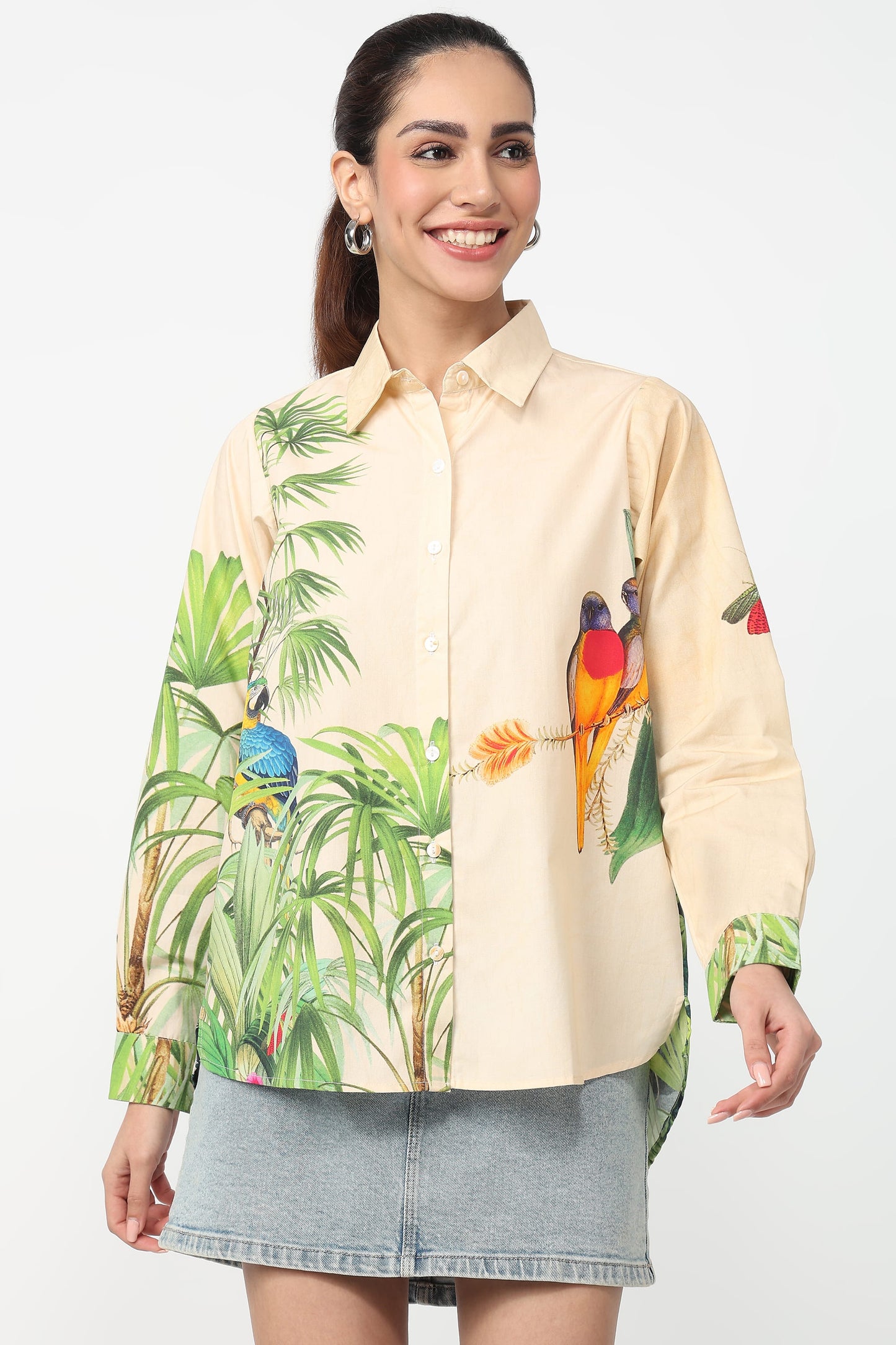 A woman modeling a long-sleeve button-up shirt with a tropical jungle print featuring green palm leaves and colorful birds, paired with a gray denim skirt against a plain white background.