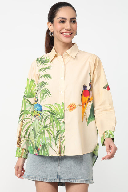 A woman modeling a long-sleeve button-up shirt with a tropical jungle print featuring green palm leaves and colorful birds, paired with a gray denim skirt against a plain white background.