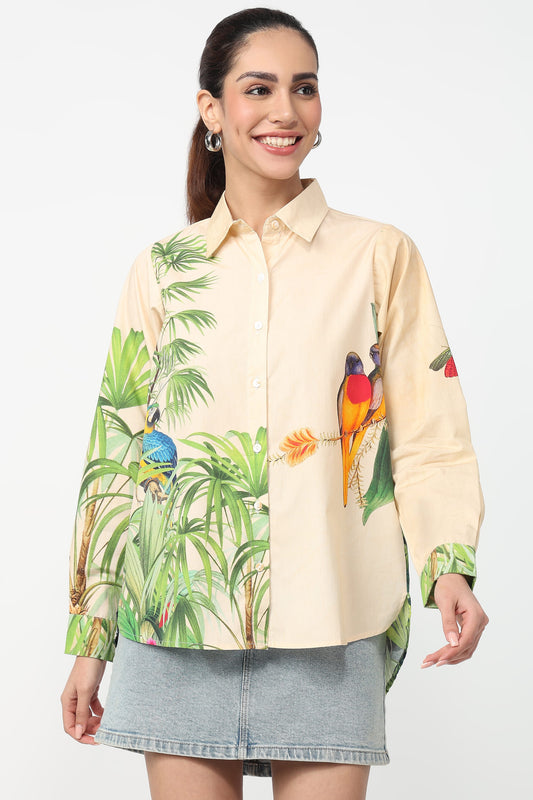 A woman modeling a long-sleeve button-up shirt with a tropical jungle print featuring green palm leaves and colorful birds, paired with a gray denim skirt against a plain white background.