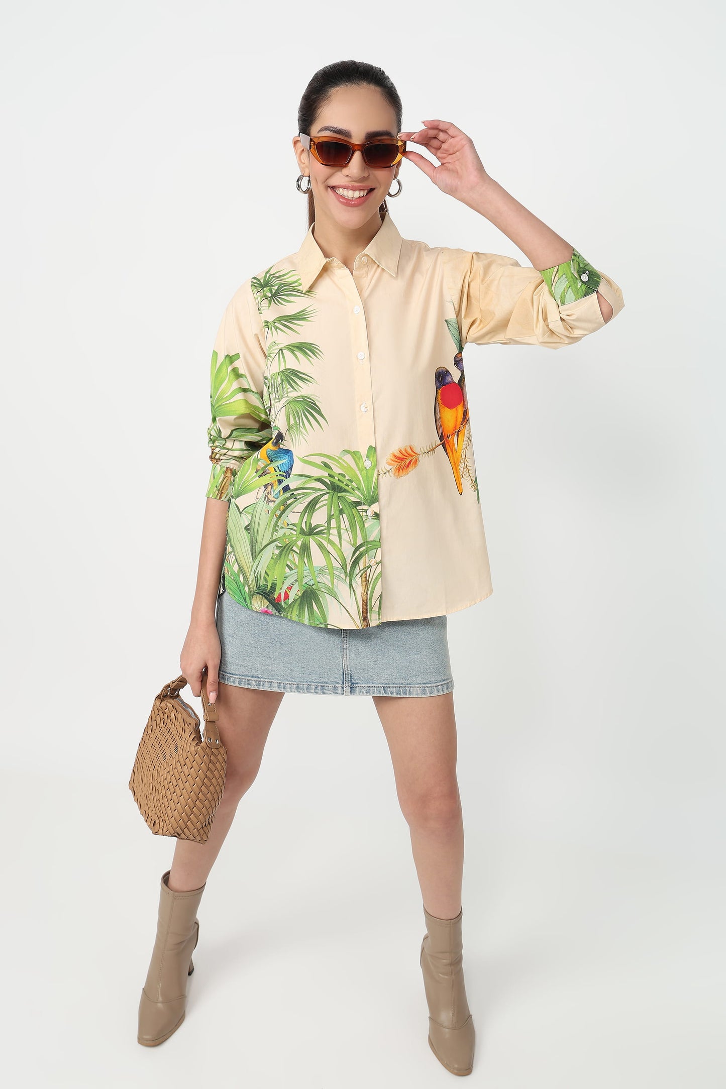 A woman wearing a beige button-up shirt with a vibrant tropical print featuring green palm leaves and colourful parrots, set against a plain light grey background.