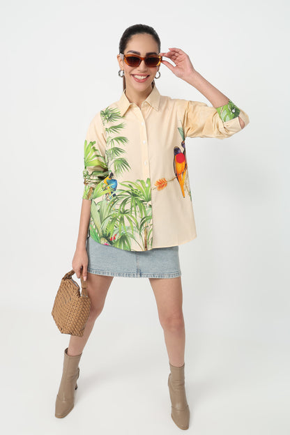 A woman wearing a beige button-up shirt with a vibrant tropical print featuring green palm leaves and colourful parrots, set against a plain light grey background.