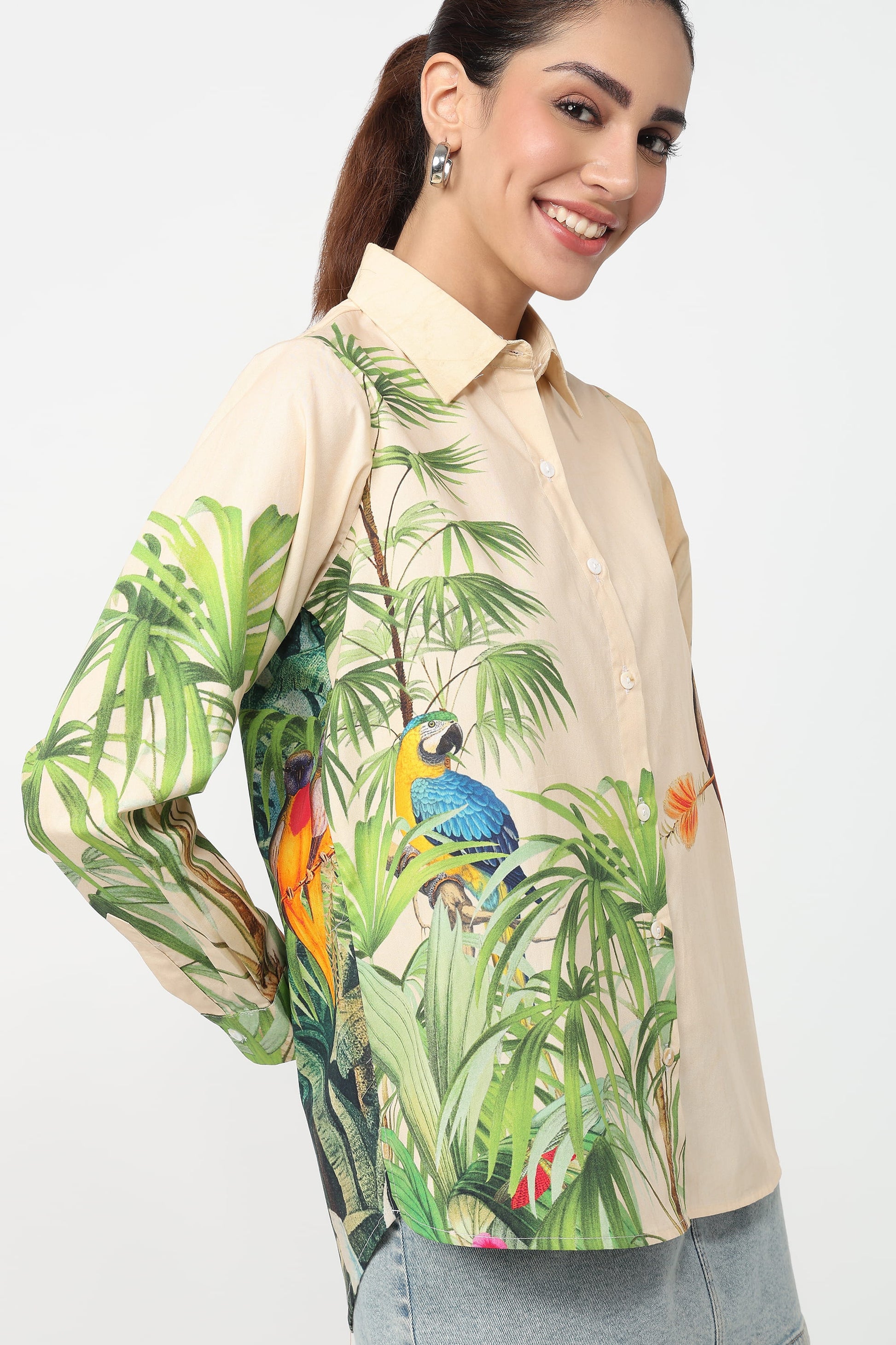 A woman wearing a beige button-up shirt with a vibrant tropical print featuring green palm leaves and colourful parrots, set against a plain light grey background.