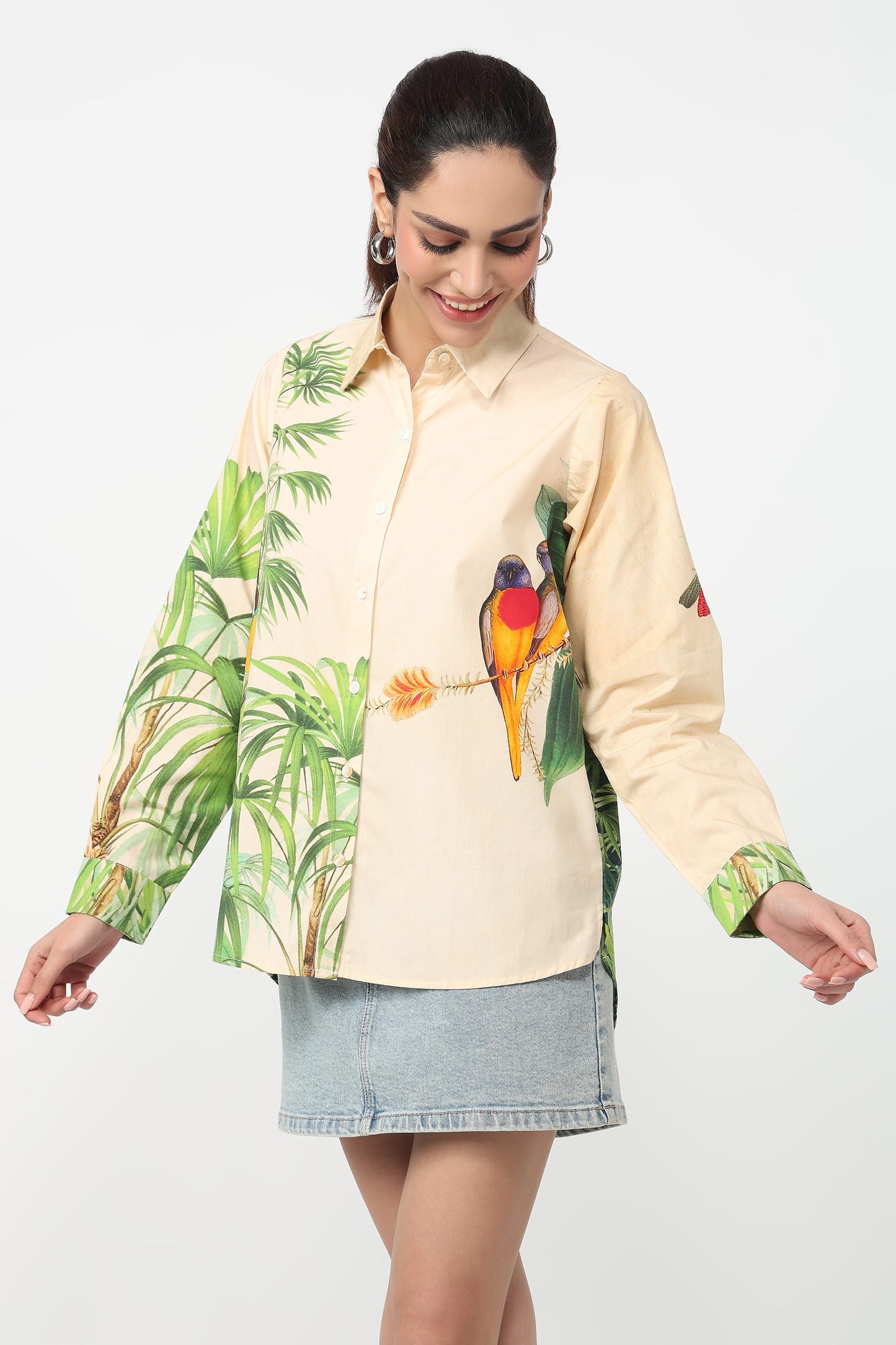 A woman wearing a beige button-up shirt with a vibrant tropical print featuring green palm leaves and colourful parrots, set against a plain light grey background.