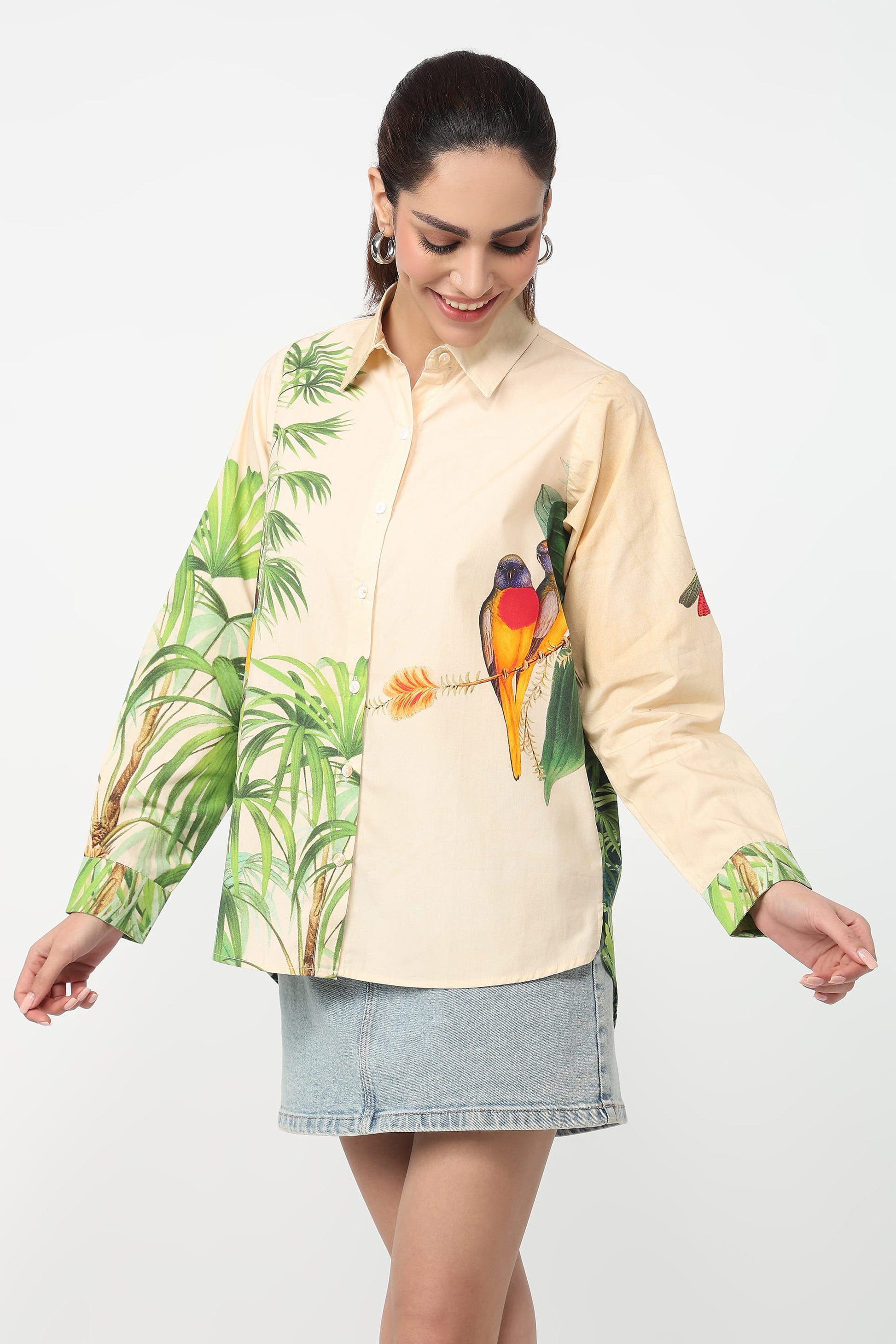 A woman wearing a beige button-up shirt with a vibrant tropical print featuring green palm leaves and colourful parrots, set against a plain light grey background.