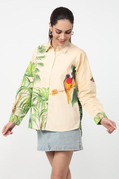 A woman wearing a beige button-up shirt with a vibrant tropical print featuring green palm leaves and colourful parrots, set against a plain light grey background.