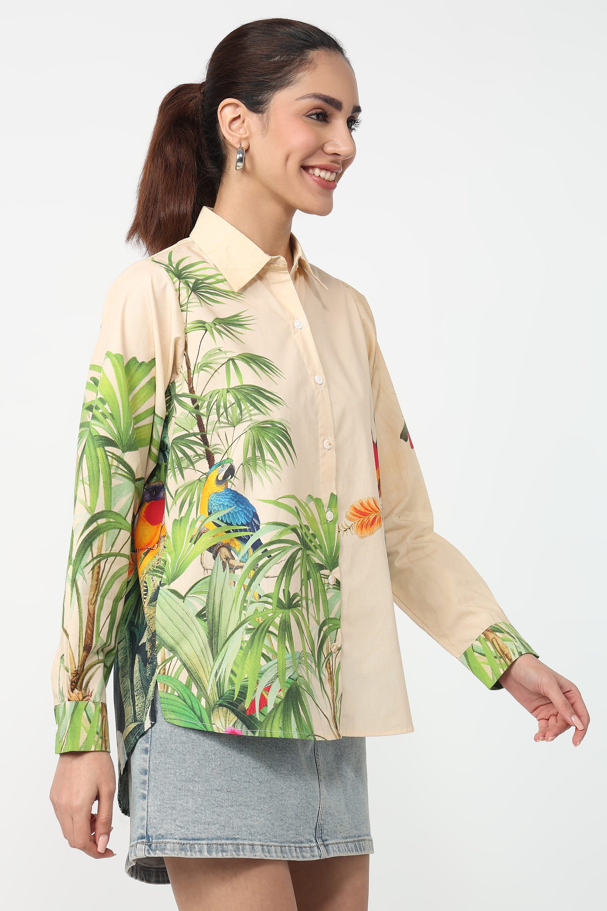 A woman wearing a beige button-up shirt with a vibrant tropical print featuring green palm leaves and colourful parrots, set against a plain light grey background.