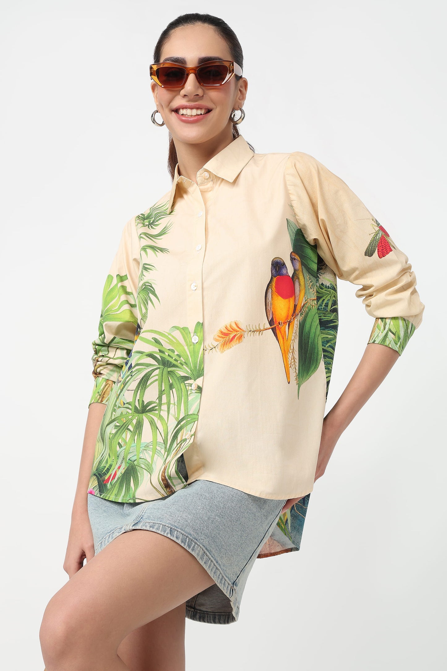 A woman wearing a beige button-up shirt with a vibrant tropical print featuring green palm leaves and colourful parrots, set against a plain light grey background.