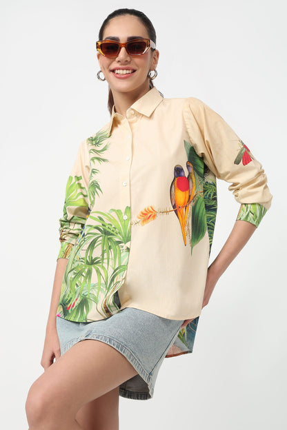 A woman wearing a beige button-up shirt with a vibrant tropical print featuring green palm leaves and colourful parrots, set against a plain light grey background.