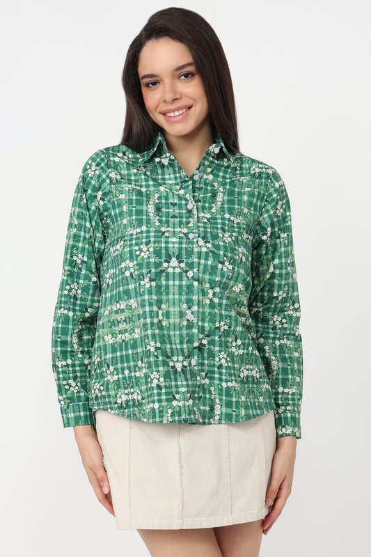 A woman modeling a green long-sleeve button-up shirt with a white floral and plaid pattern, paired with a cream-colored pleated mini skirt, against a plain white studio background.