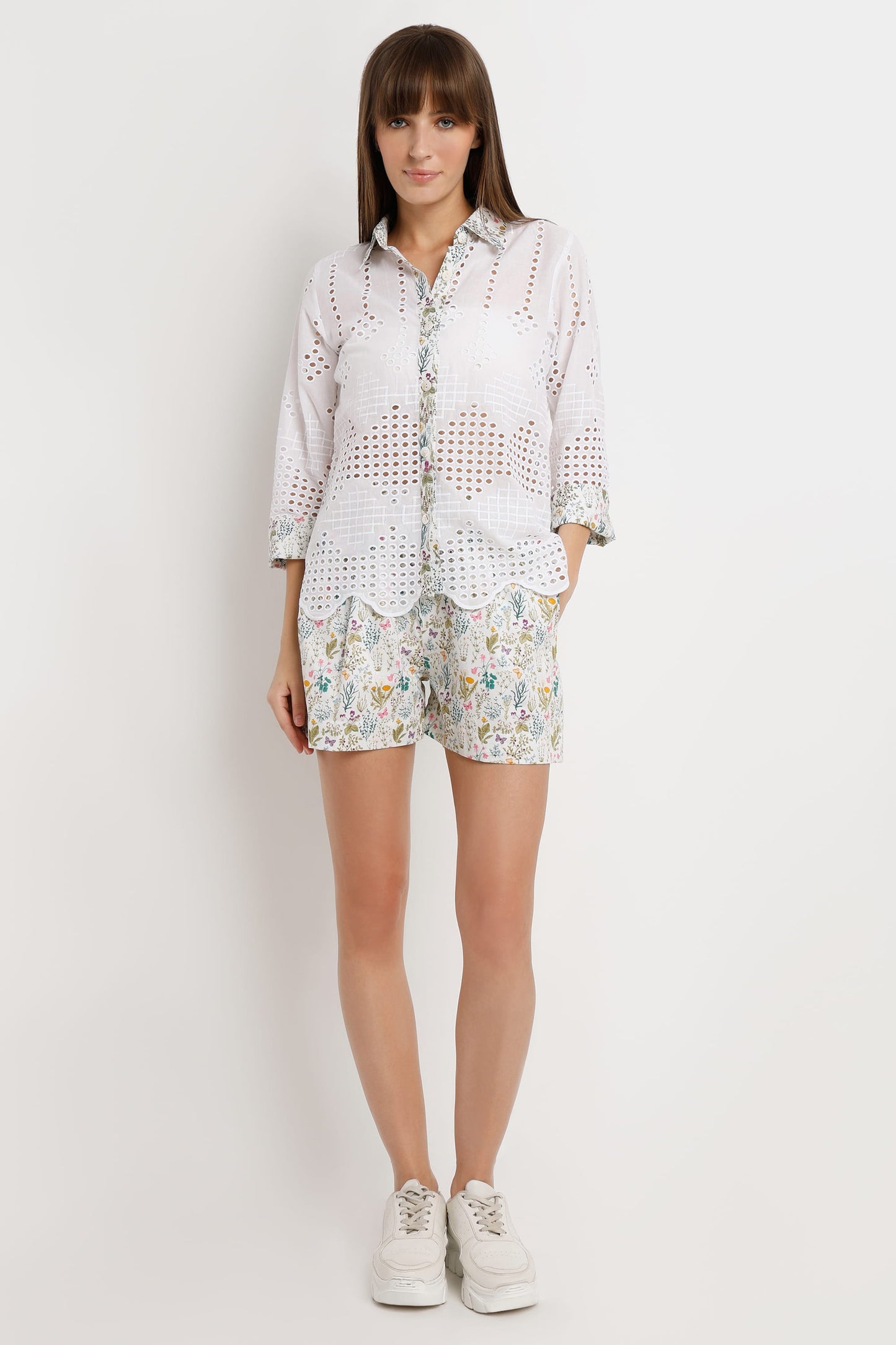 A woman modeling a white eyelet button-up shirt and matching floral print shorts against a plain light gray studio background.