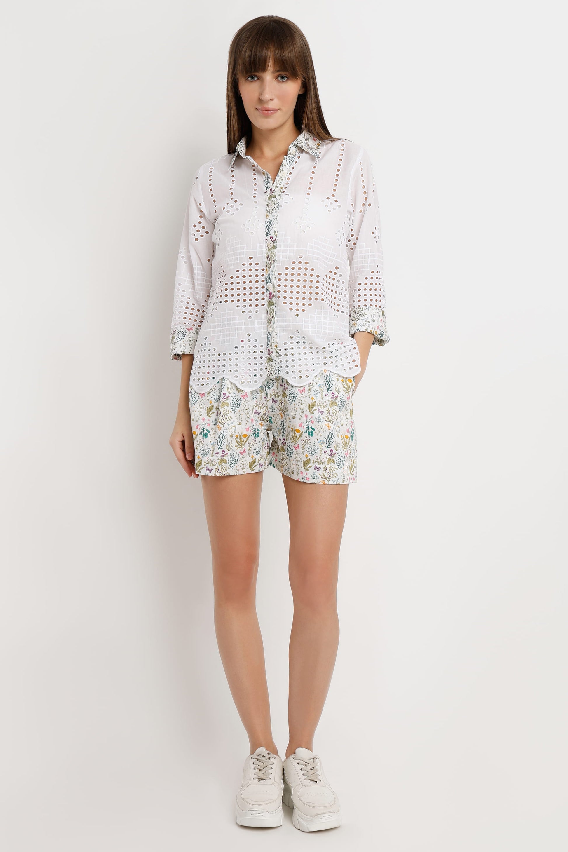A woman modeling a white eyelet button-up shirt and matching floral print shorts against a plain light gray studio background.