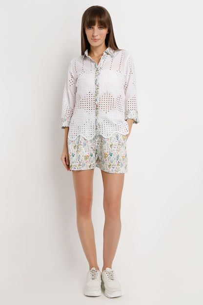 A woman modeling a white eyelet button-up shirt and matching floral print shorts against a plain light gray studio background.