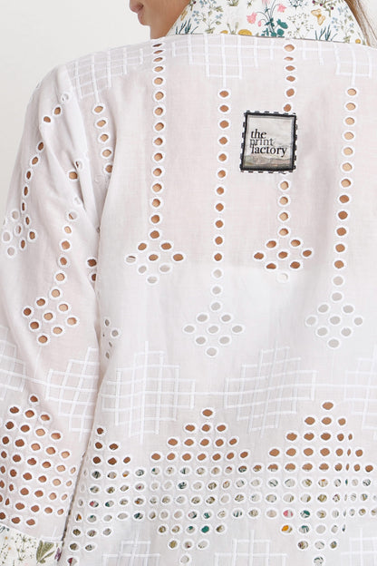 Close-up of a white eyelet embroidered jacket with geometric cutout patterns, featuring a black and white 'the tiny factory' brand patch on the back.