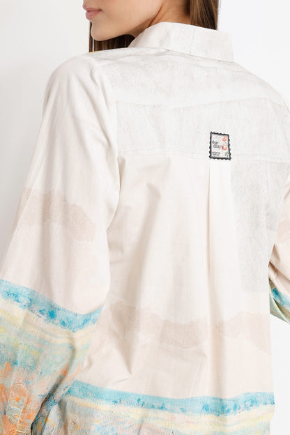 Close-up back view of a model wearing a cream-colored shirt with a small square patch on the upper back and a blue and orange patterned border near the bottom.