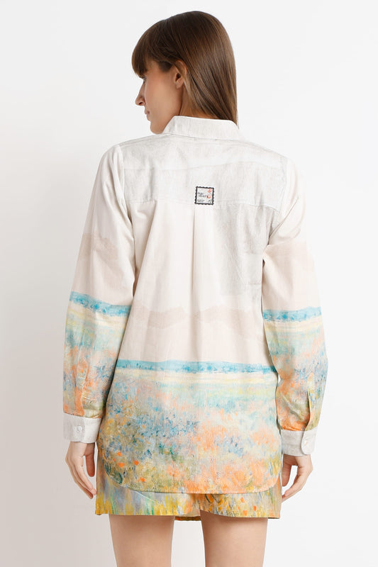 A woman shown from the back wearing a light beige button-up shirt dress with a colorful abstract watercolor-style print in blue, yellow, and orange on the sleeves and lower hem.