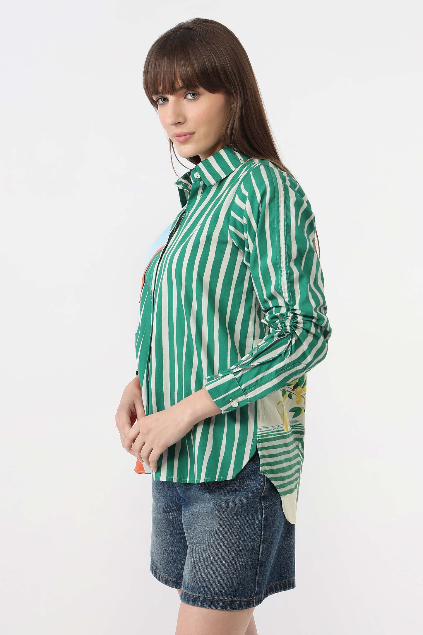 A woman wearing a green and white vertically striped button-up shirt with rolled-up sleeves and blue denim shorts, photographed against a plain white studio background.