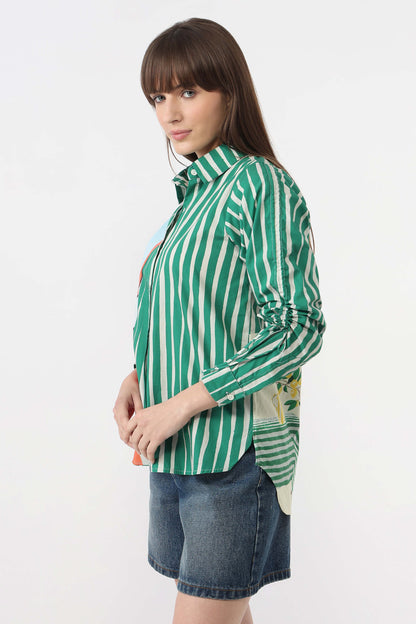A woman wearing a green and white vertically striped button-up shirt with rolled-up sleeves and blue denim shorts, photographed against a plain white studio background.