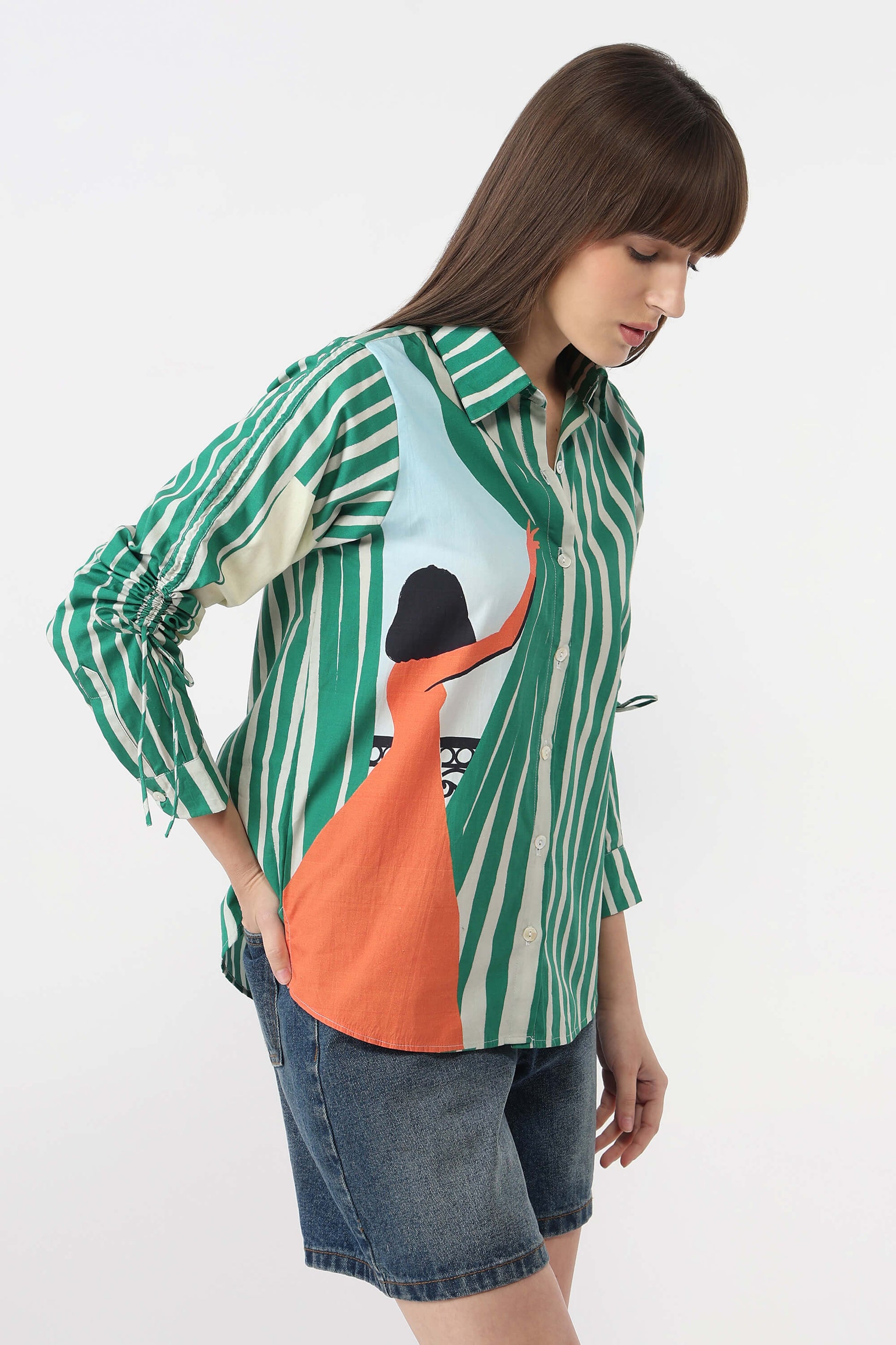 A woman wearing a green and white vertically striped button-up shirt with a large orange and black graphic print on the front, paired with denim shorts, against a plain white studio background.