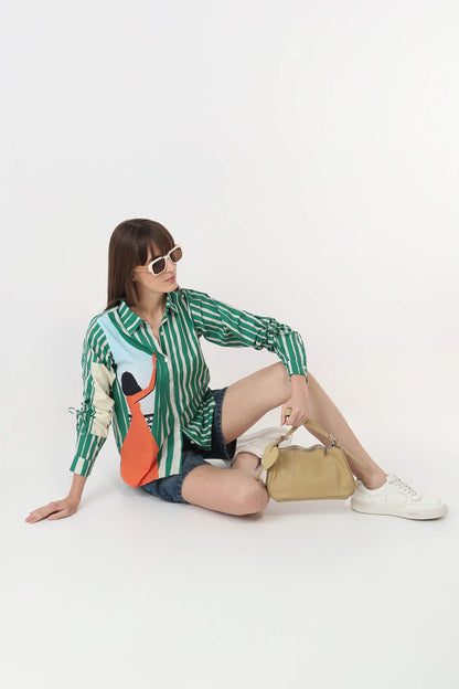 A woman wearing a green and white striped button-up shirt with a colorful graphic print, denim shorts, white sneakers, and sunglasses sits on a white studio floor next to a beige handbag.