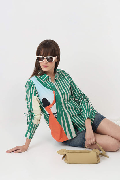 A woman wearing a green and white striped button-up shirt with a colorful graphic print, denim shorts, and white sunglasses, sitting on a white studio floor next to a beige handbag.