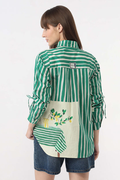 A woman shown from the back wearing a green and white striped shirt with a large cream-colored patch pocket featuring a lemon illustration, paired with denim shorts, against a plain white background.