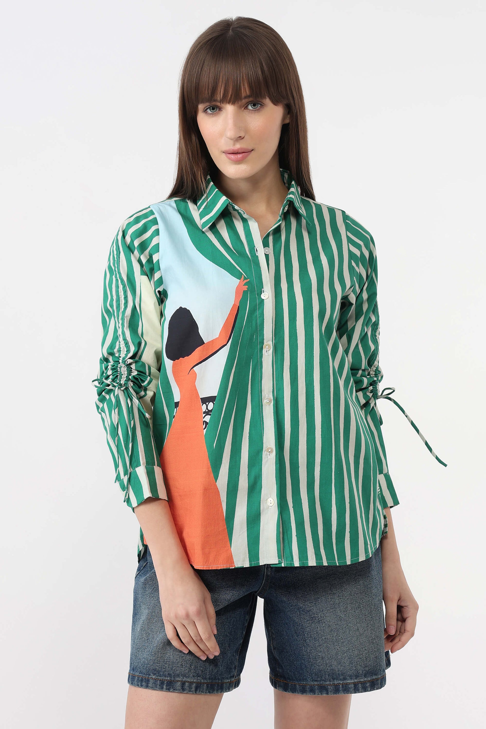 A woman modeling a green and white vertically striped button-up shirt with a large graphic print on the front, paired with denim shorts, against a plain white studio background.