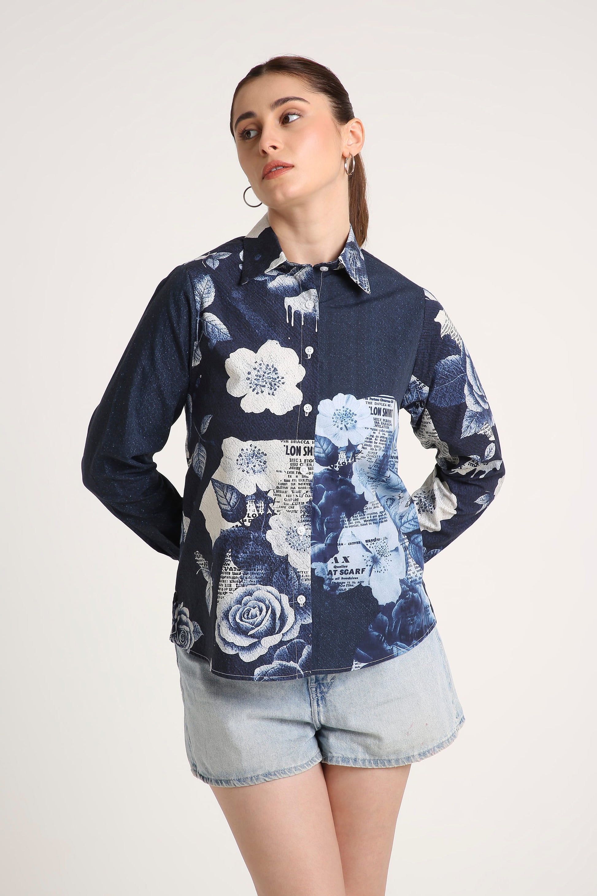 A woman wearing a long-sleeve button-up shirt with a navy blue base and a white floral print, paired with light denim shorts, against a plain light background.