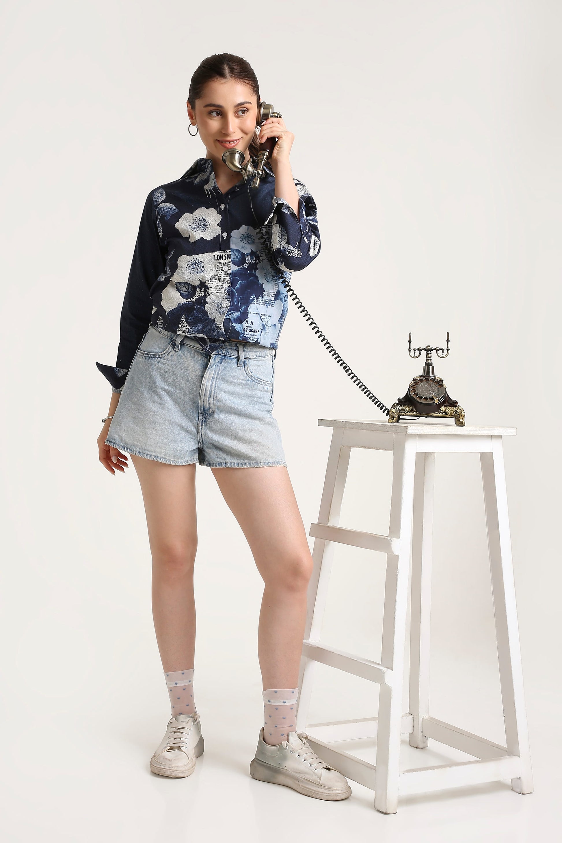 A woman stands in a studio wearing a navy floral long-sleeve shirt, light denim shorts, white socks, and white sneakers, holding a vintage black rotary telephone to her ear. A matching white stool with the telephone sits beside her against a plain light background.