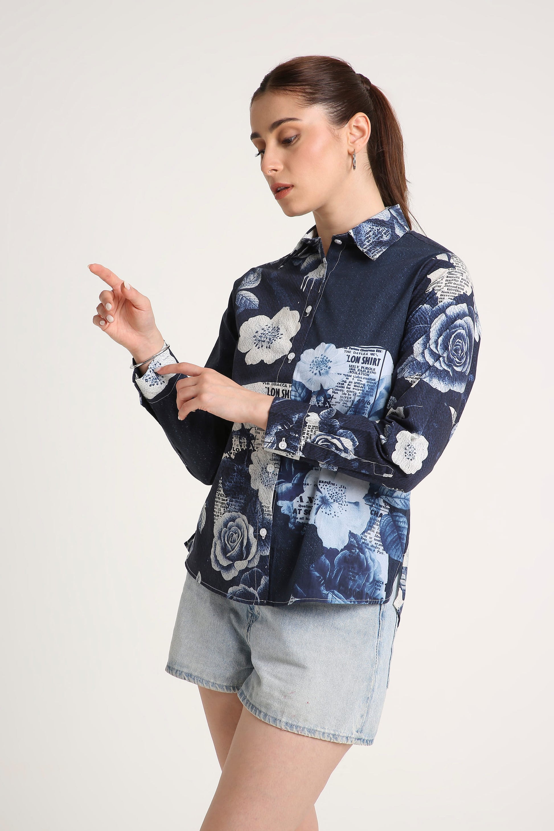 A woman wearing a navy blue button-up shirt with a white and light blue floral print, paired with light-wash denim shorts, against a plain off-white studio background.