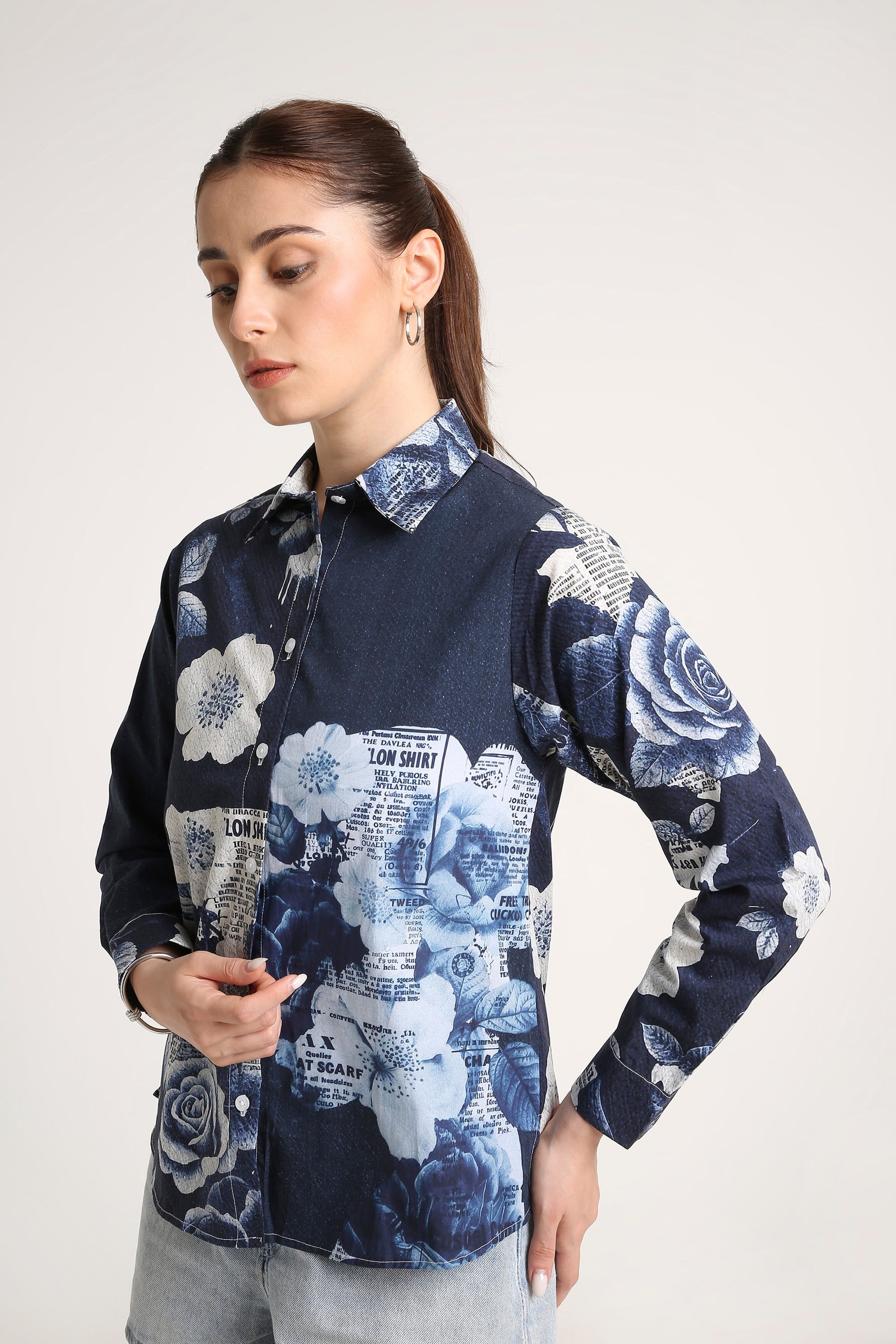 A woman wearing a long-sleeve button-up shirt with a navy blue base, featuring a floral rose print on the sleeves and a blue-and-white collage print on the front, paired with light gray jeans against a plain light gray studio background.