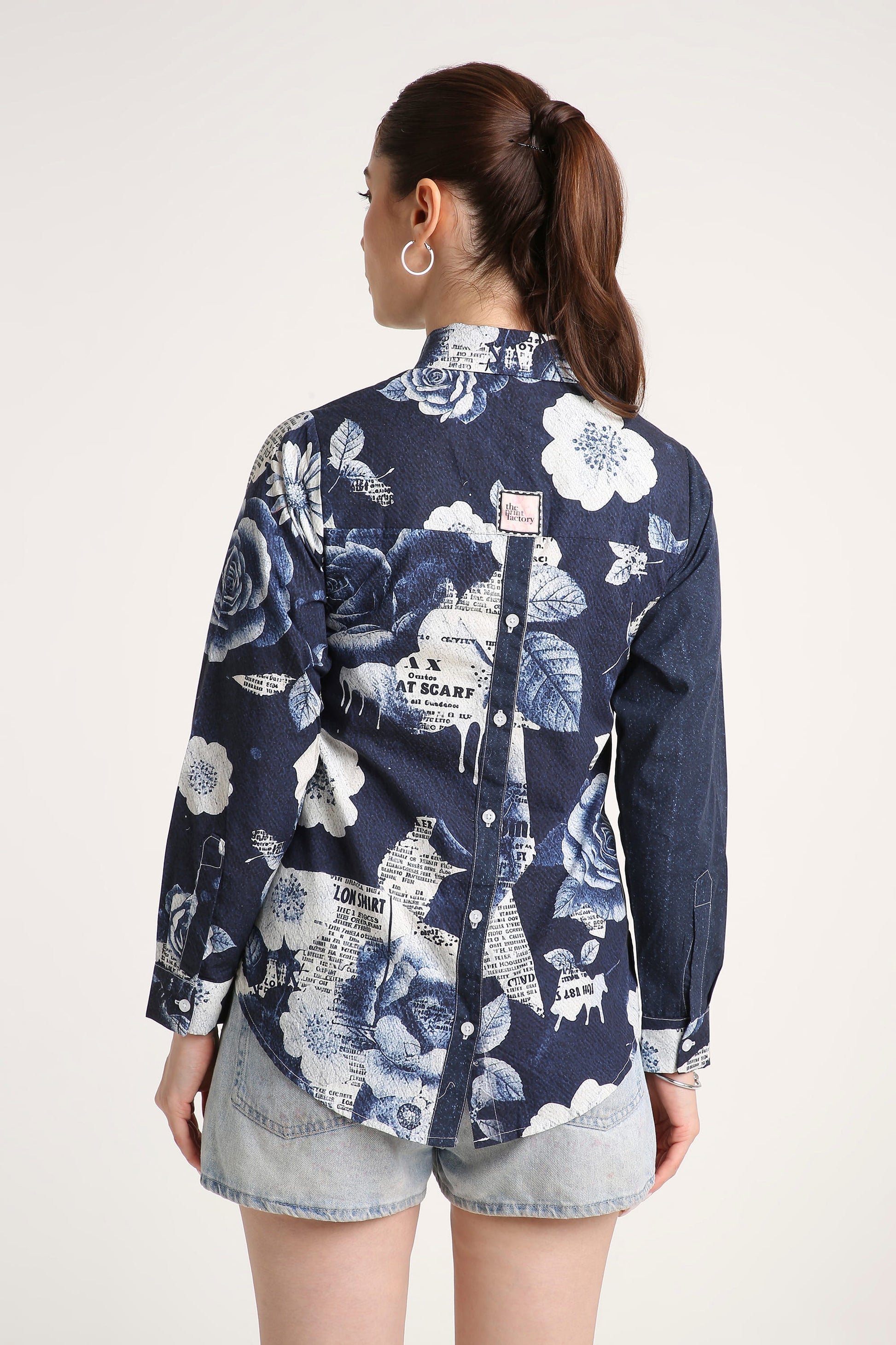 A woman shown from the back wearing a navy blue long-sleeve button-up shirt with a white floral and newspaper print, paired with denim shorts, against a plain light background.