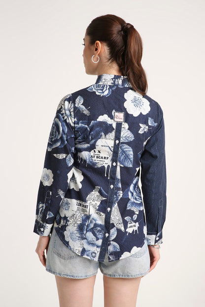 A woman shown from the back wearing a navy blue long-sleeve button-up shirt with a white floral and newspaper print, paired with denim shorts, against a plain light background.