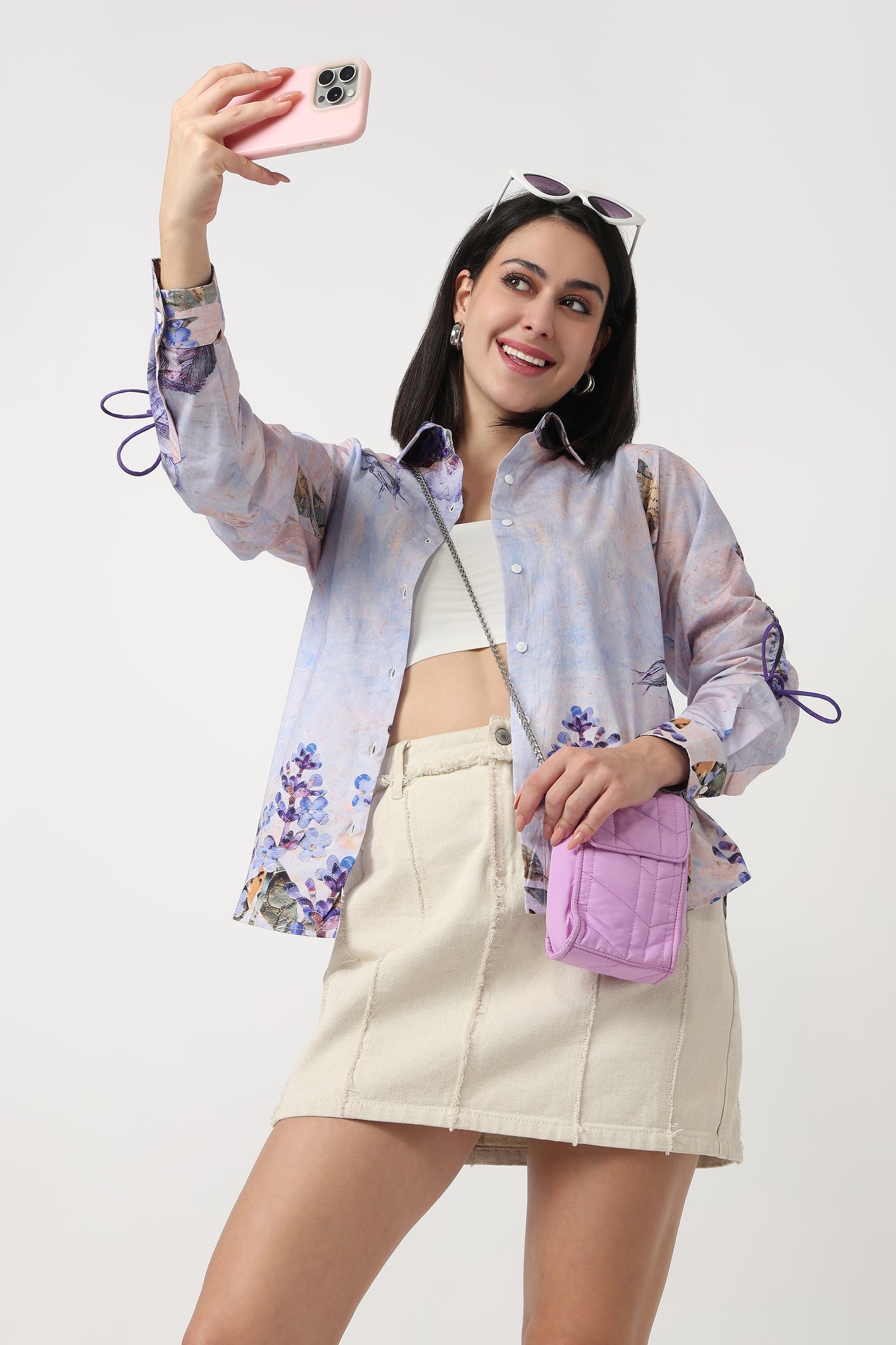 Woman taking a selfie wearing a floral shirt, white top, beige shorts, and a pink bag on a white background