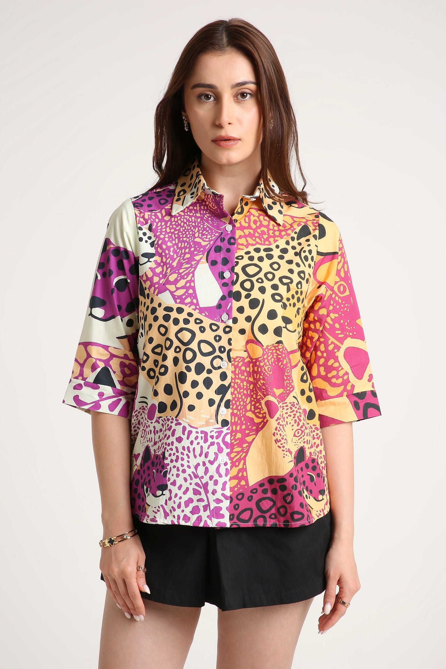 A woman wearing a short-sleeve button-up shirt with a vibrant multicolour patchwork print featuring leopard and cheetah patterns in pink, purple, yellow, and black, paired with black shorts against a light grey background.