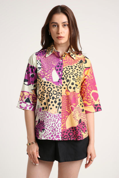 A woman wearing a short-sleeve button-up shirt with a vibrant multicolour patchwork print featuring leopard and cheetah patterns in pink, purple, yellow, and black, paired with black shorts against a light grey background.