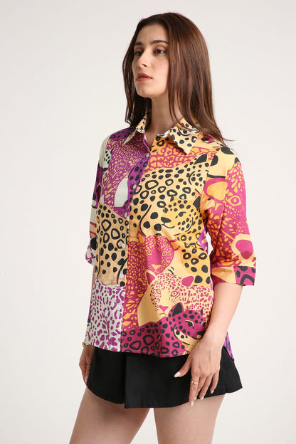 A woman wearing a short-sleeve button-up shirt with a vibrant multicolour abstract print featuring leopard and floral motifs in pink, yellow, purple, and black, paired with black shorts against a light grey background.