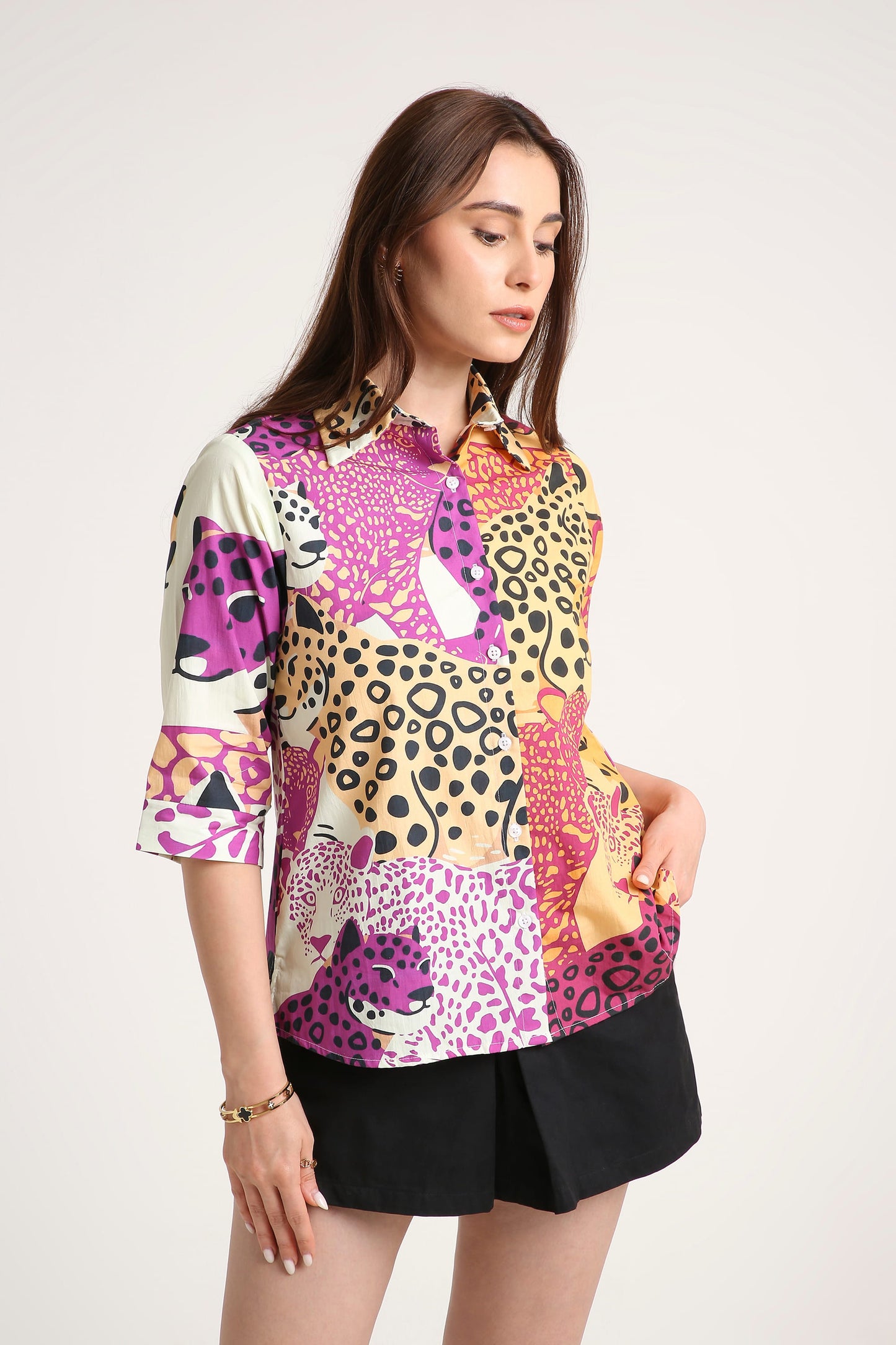 A woman wearing a short-sleeve button-up shirt with a vibrant multicolour animal print pattern featuring pink, purple, black, and beige leopard and cheetah motifs, paired with black shorts against a plain light background.